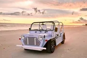 A light blue beach buggy is parked on the sand with a beautiful ocean sunset in the background.
