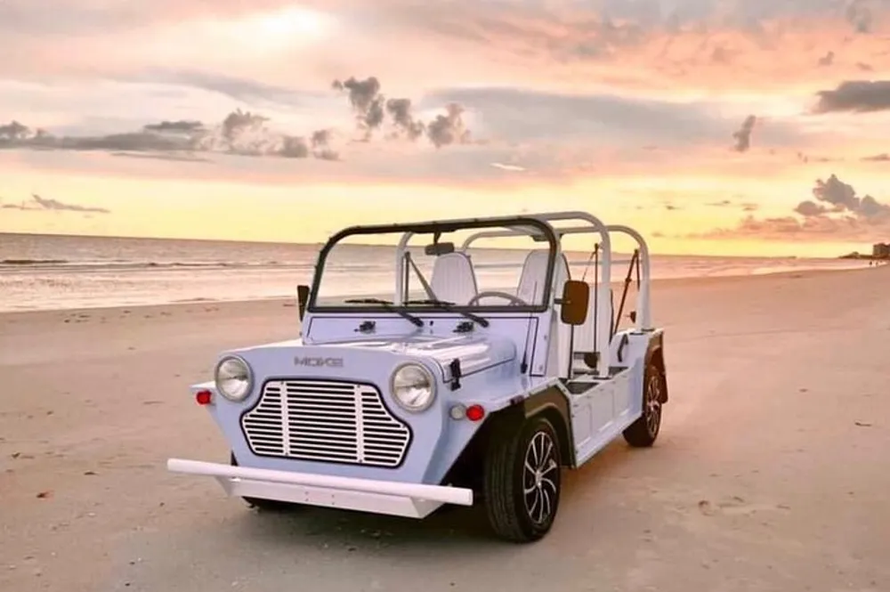 A light blue beach buggy is parked on the sand with a beautiful ocean sunset in the background