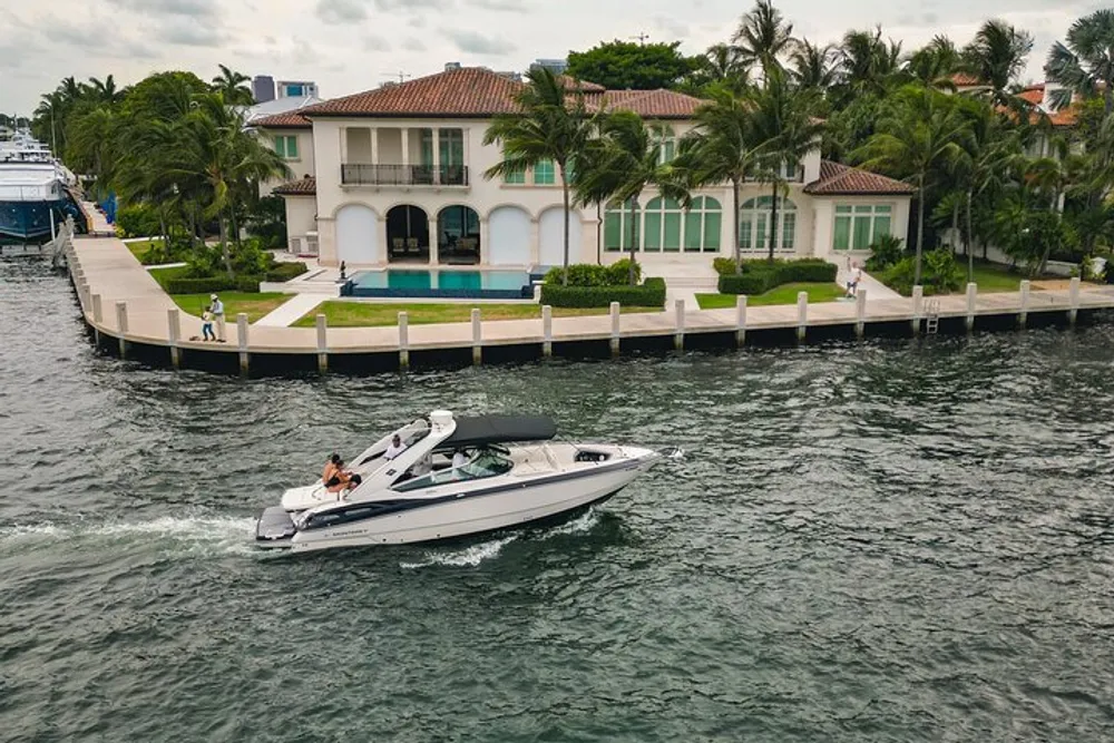 A motorboat cruises past a luxurious waterfront home with a person standing on the dock