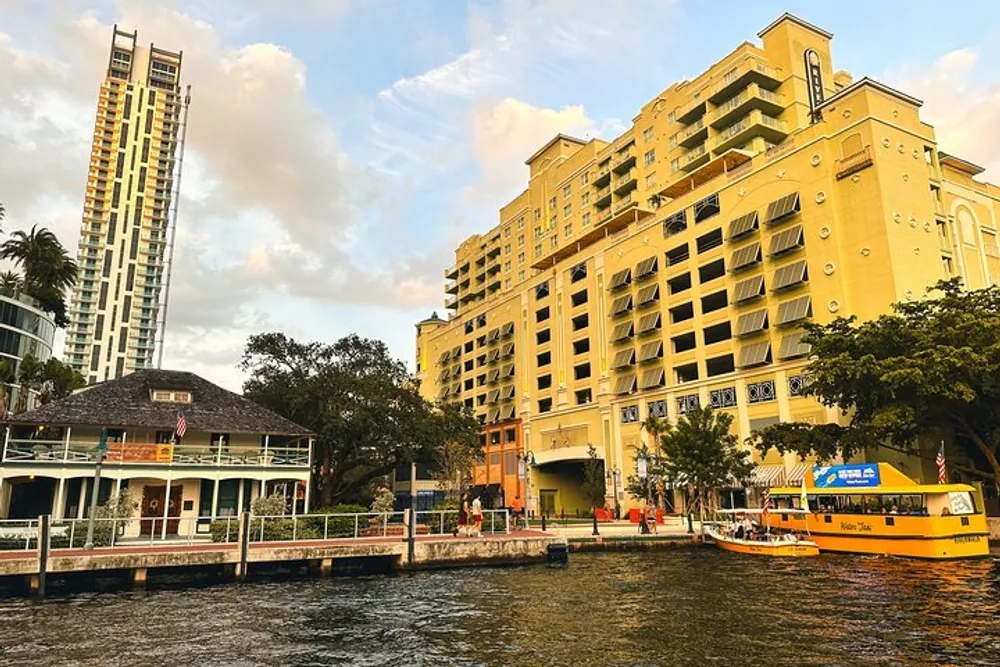The image captures a waterfront scene with a yellow water taxi docked near a pavilion flanked by a mix of modern and classical architecture under a partly cloudy sky