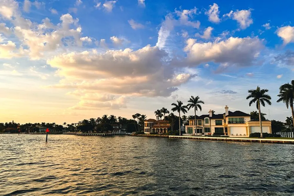 The image shows opulent waterfront homes under a dynamic sky at sunset with silhouetted palm trees adding to the tranquil tropical scenery