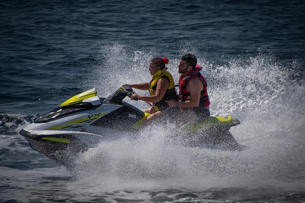 Two people wearing life jackets are riding a jet ski on the water creating a spray of water as they go