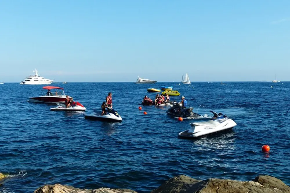 The image depicts a bustling aquatic scene with various people engaged in recreational activities such as riding jet skis and a speedboat with larger yachts and sailboats visible in the distance all set against a deep blue sea and clear sky