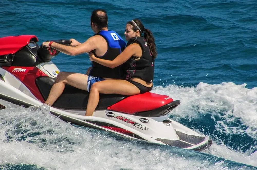 Two people in life jackets are riding a red and white jet ski across sparkling blue water