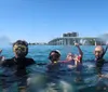 Four people wearing snorkeling gear are making the OK hand signal while floating in clear water with a bridge and buildings in the background