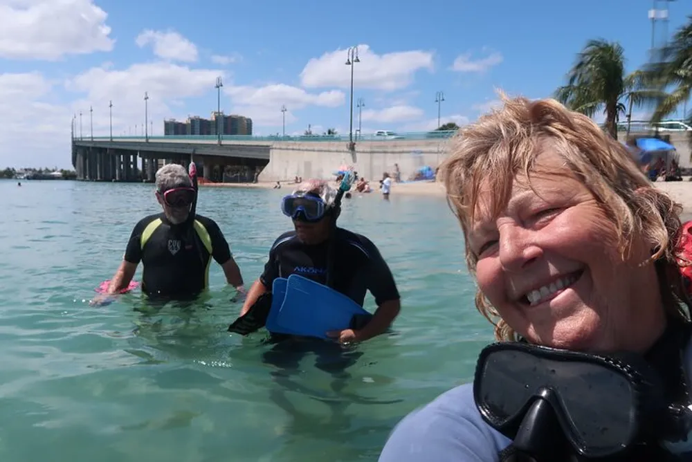 A person takes a selfie with two others wearing snorkeling gear in shallow beach waters near a pier