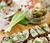 This image features an appetizing arrangement of open sandwiches with various toppings surrounded by fresh ingredients and pickles on a wooden surface