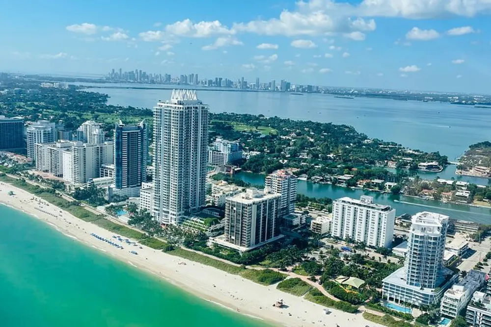 This image shows a sunny aerial view of high-rise buildings along a beach with a clear blue ocean on one side and a city skyline in the distance