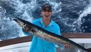 A person is holding a large fish with a big smile on their face, standing on a boat with the ocean in the background.