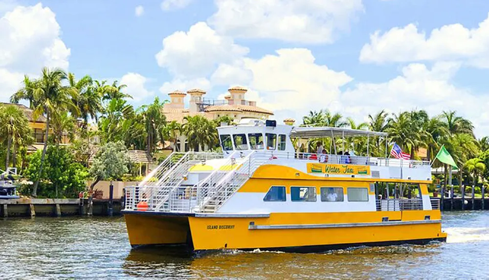 A yellow and white water taxi boat is cruising along a waterway with tropical palm trees and an elegant mansion in the background
