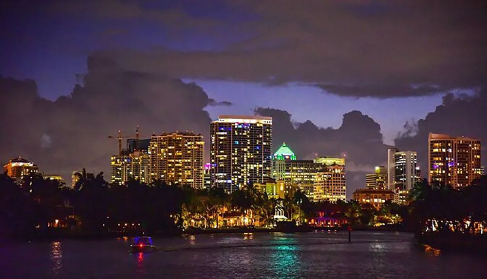The image captures a nighttime view of a city skyline illuminated with lights reflecting on the water with a dramatic cloud-filled sky in the background