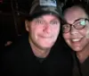Two people are smiling for a selfie in a dimly lit environment with one person wearing a baseball cap and the other wearing glasses