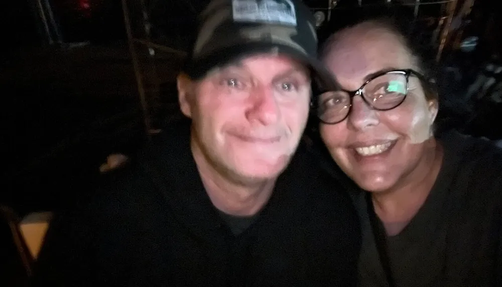 Two people are smiling for a selfie in a dimly lit environment with one person wearing a baseball cap and the other wearing glasses