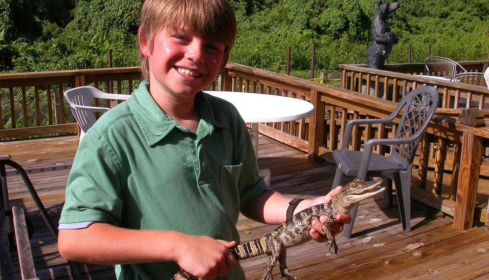 A smiling boy is holding a small alligator on a sunny deck with outdoor furniture in the background