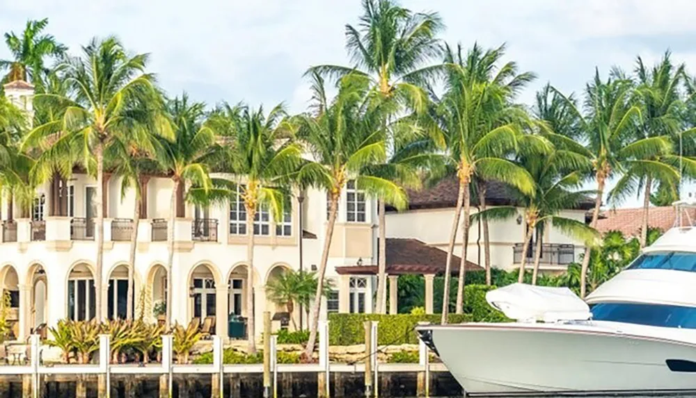 A luxury waterfront house with palm trees is complemented by a moored white yacht on a sunny day