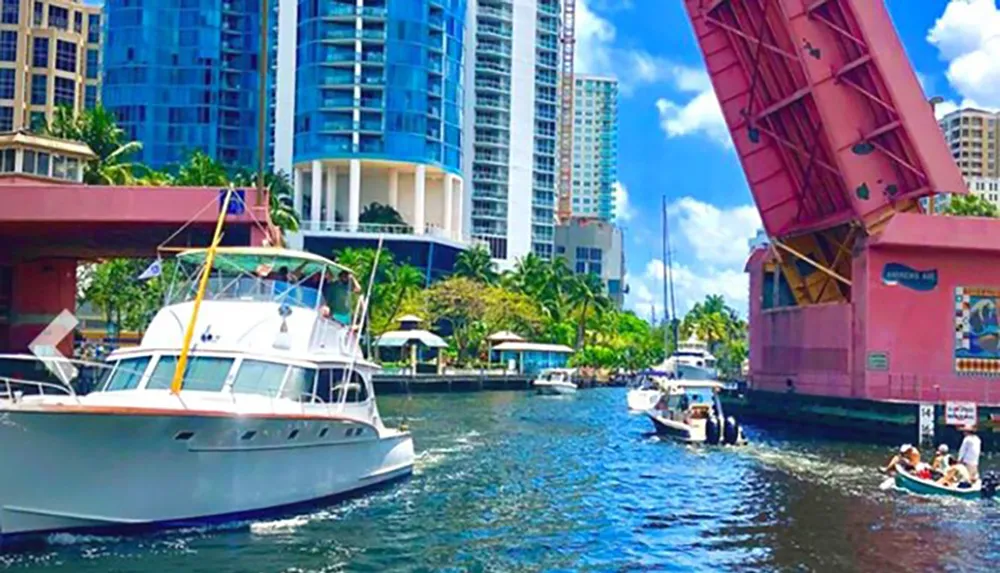 A yacht passes under a raised drawbridge on a sunlit waterway flanked by high-rise buildings and lush greenery
