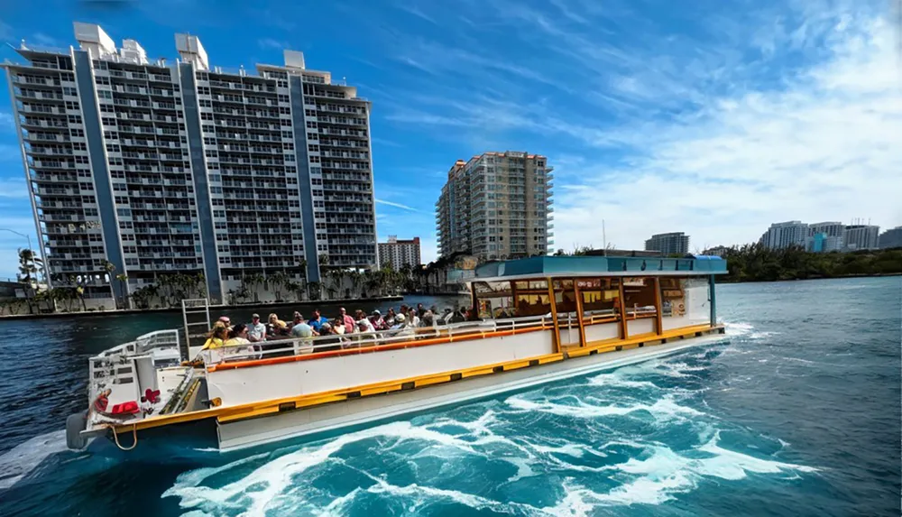 A water taxi full of passengers is cruising along a waterway with high-rise buildings in the background under a vivid blue sky