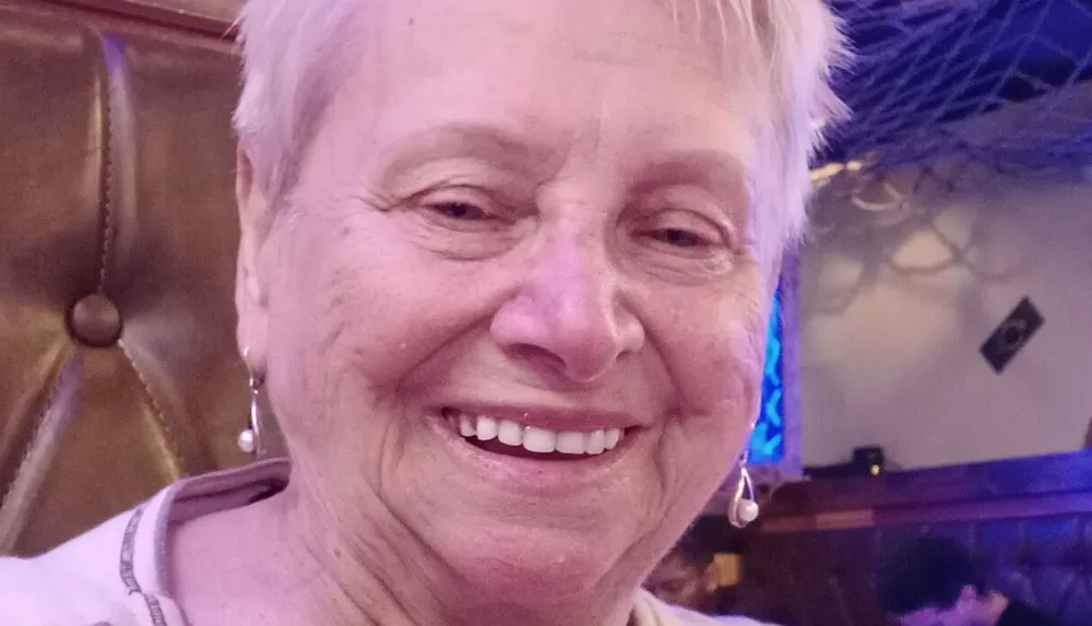 A smiling person with short hair and earrings is posing for a close-up photo in a room with a warm ambiance