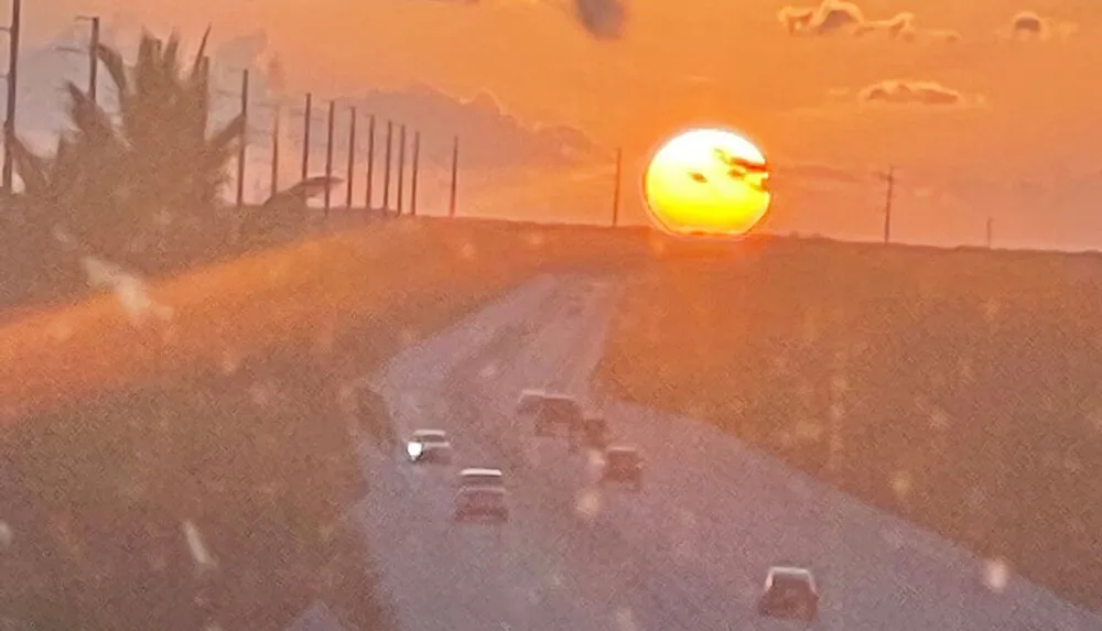 The image captures a dramatic large setting sun in a hazy orange sky casting a warm glow over a road with several cars driving flanked by electricity poles and a silhouette of vegetation