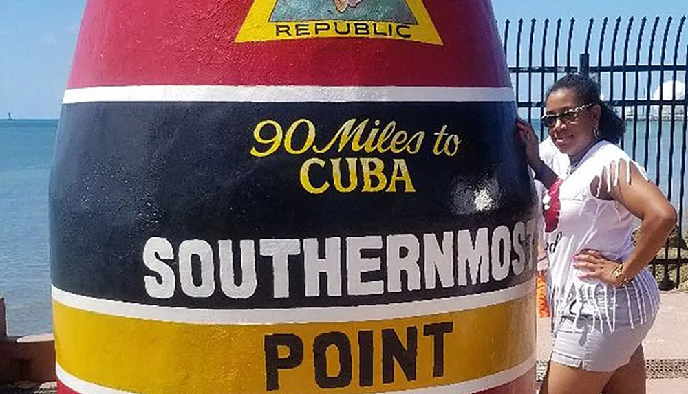 A person is posing next to the iconic Southernmost Point buoy in Key West Florida with the text 90 Miles to Cuba