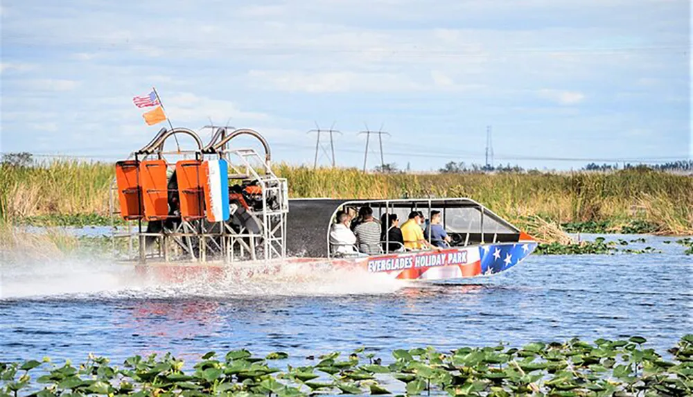 An airboat carrying passengers glides across the water amidst the greenery of a wetland with an American flag displayed at the rear