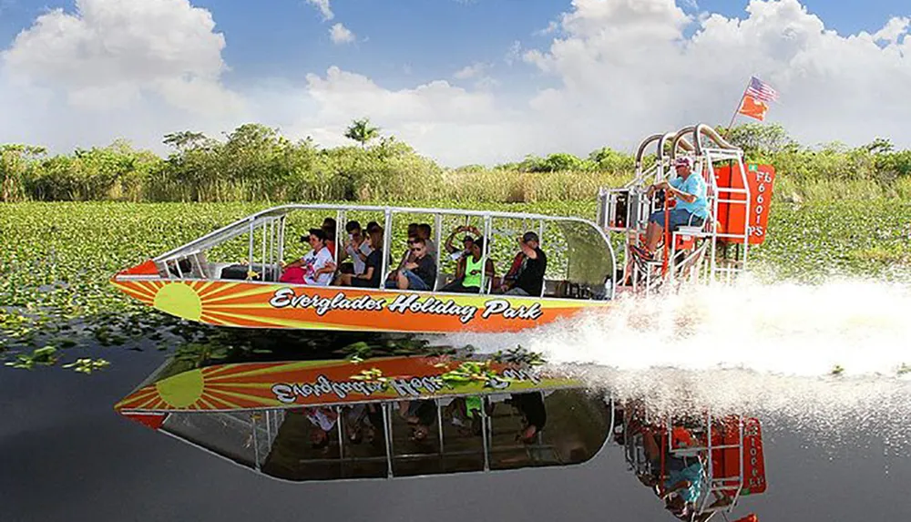 This image shows passengers enjoying a ride on an airboat skimming over the water in the Everglades with lush greenery in the background and the boats reflection visible on the waters surface