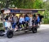 A group of people are socializing and pedaling together on a multi-passenger pedal-powered vehicle commonly known as a party bike or pedal pub on a suburban street