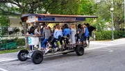 A group of people are socializing and pedaling together on a multi-passenger pedal-powered vehicle, commonly known as a party bike or pedal pub, on a suburban street.