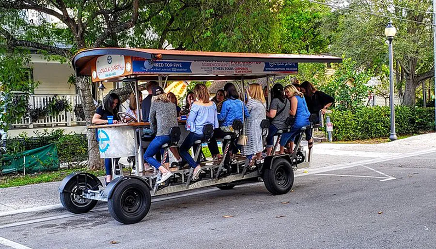 A group of people are socializing and pedaling together on a multi-passenger pedal-powered vehicle, commonly known as a party bike or pedal pub, on a suburban street.