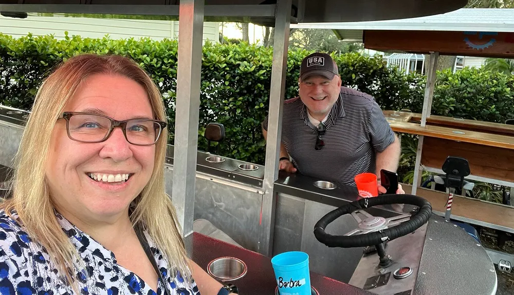 A woman is taking a selfie with a smiling man in the background who is standing behind what appears to be a mobile bar setup with personal cups in view indicating a casual and social setting