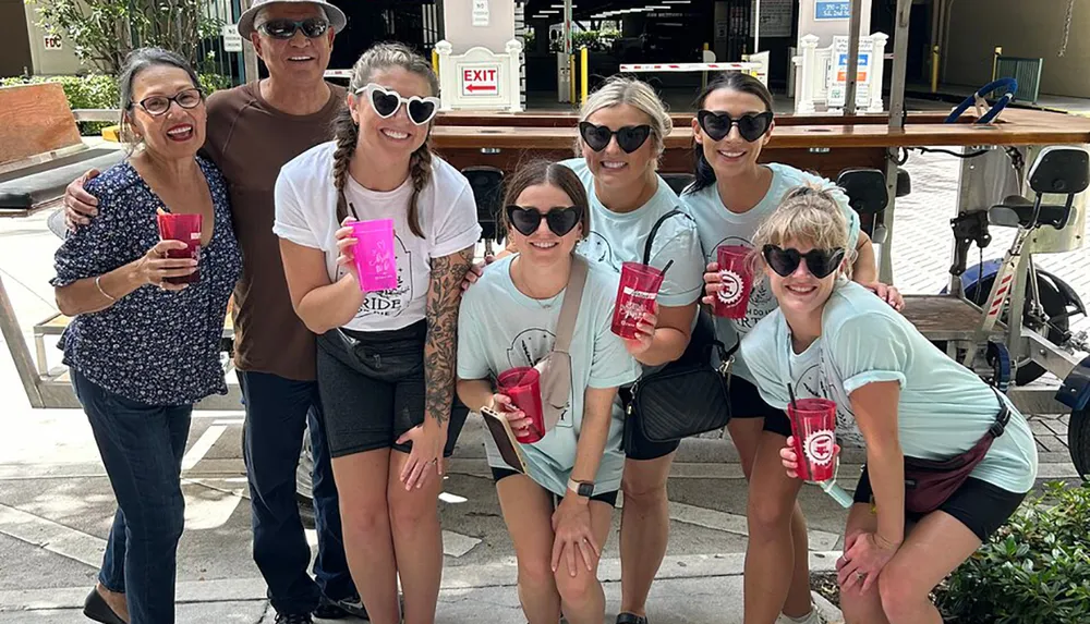 A group of seven people some wearing matching t-shirts are smiling for a photo while holding red and pink cups possibly during a group outing or celebration
