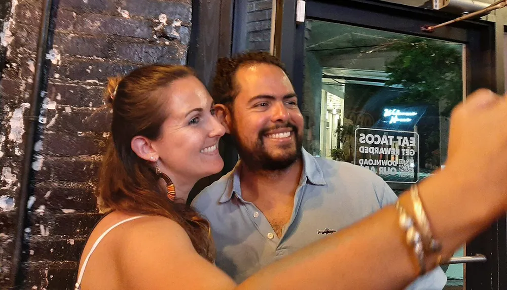 A smiling man and woman take a selfie together in a lively urban environment at night