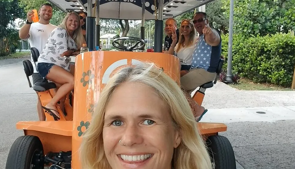 A group of happy people are enjoying a ride on a quirky orange multi-passenger pedal-powered vehicle with one person in the foreground taking a selfie