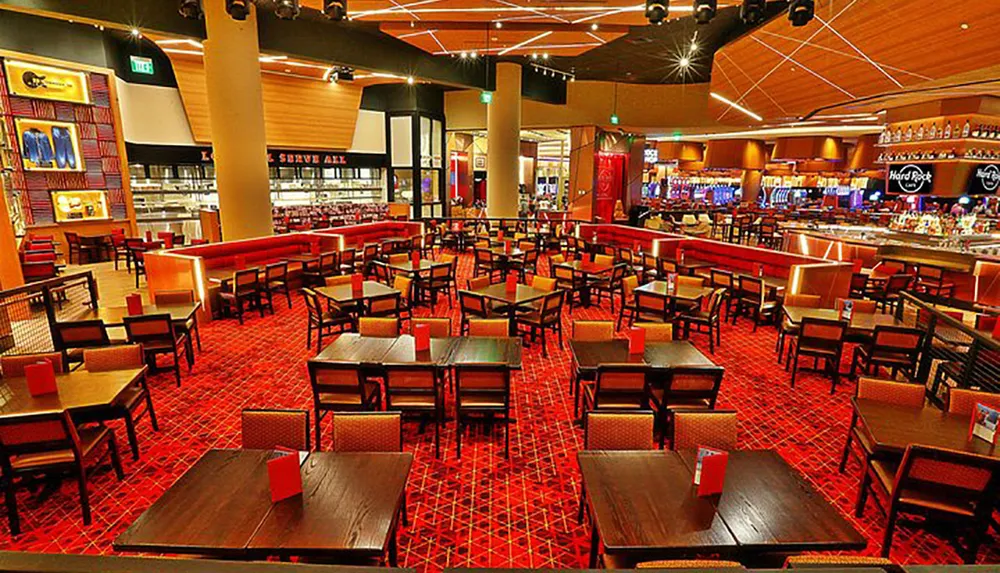 The image shows an expansive brightly-lit interior of a restaurant or dining area with numerous empty tables and chairs on a patterned red carpet with a bar in the background