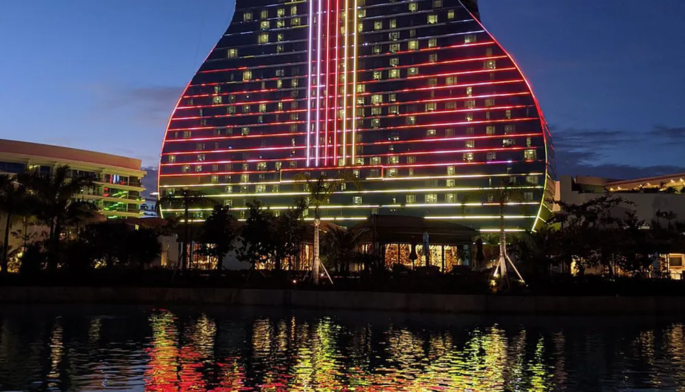 The image shows a modern illuminated building with neon lights reflecting on the waters surface at dusk