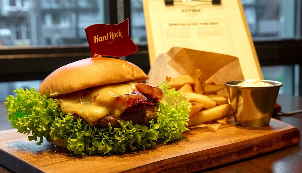 A juicy cheeseburger with bacon lettuce and a tomato on a wooden serving board is accompanied by a side of fries and dip against the backdrop of a casual diner setting