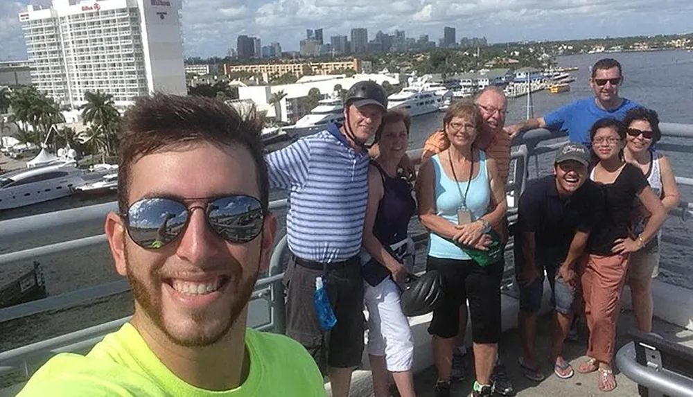 A group of smiling people are posing for a selfie with a scenic waterfront and city skyline in the background
