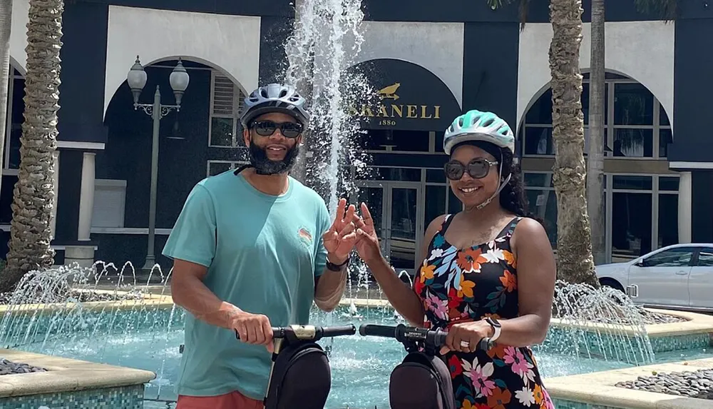Two people wearing helmets and sunglasses are standing smiling with bicycles in front of a fountain and a boutique