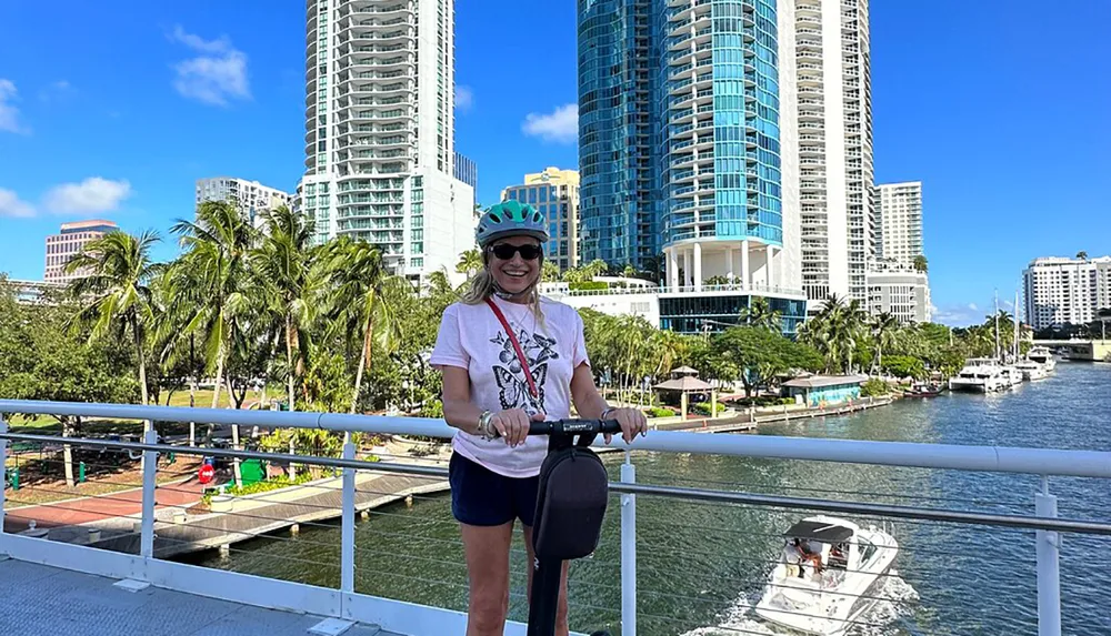 A person is smiling while standing on a bridge with a scooter wearing a helmet and sunglasses with a picturesque backdrop of a river boats and high-rise buildings under a clear blue sky