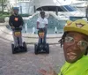 Three people are posing with two on Segways and one taking a selfie all smiling and having a good time near a fountain and yacht