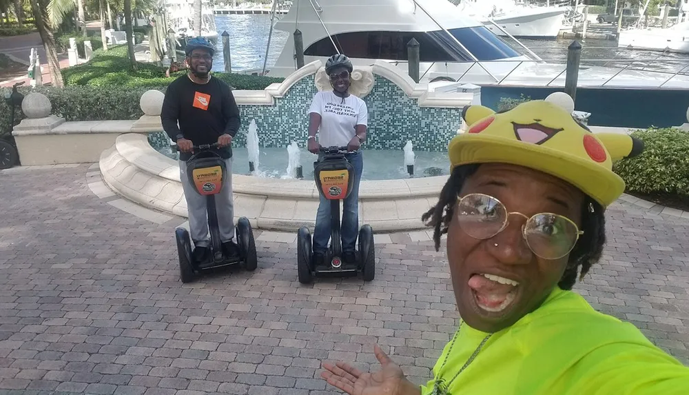 Three people are posing with two on Segways and one taking a selfie all smiling and having a good time near a fountain and yacht