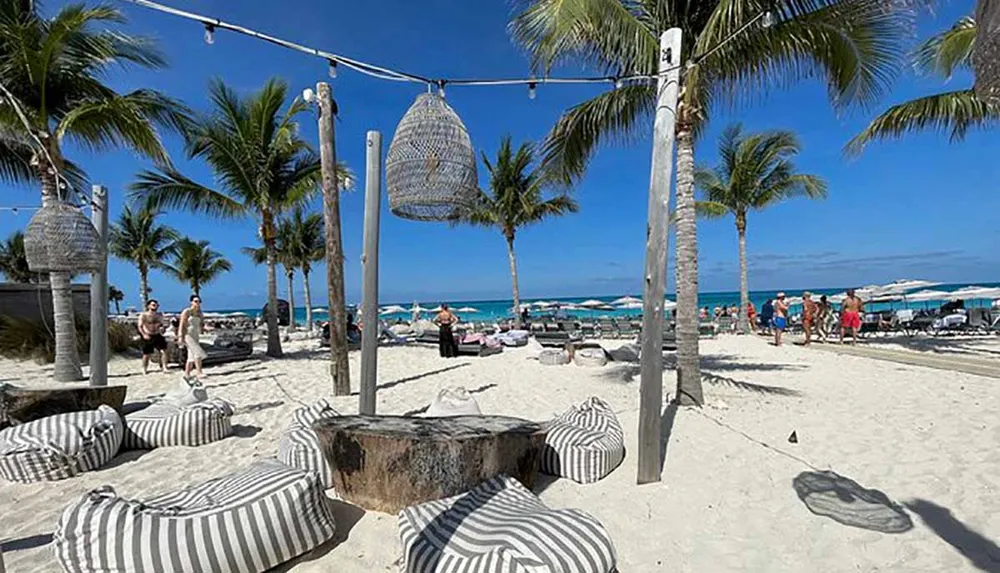 This image shows a sunny beach scene with striped beanbag chairs palm trees hanging basket chairs and people enjoying the day with the ocean in the background