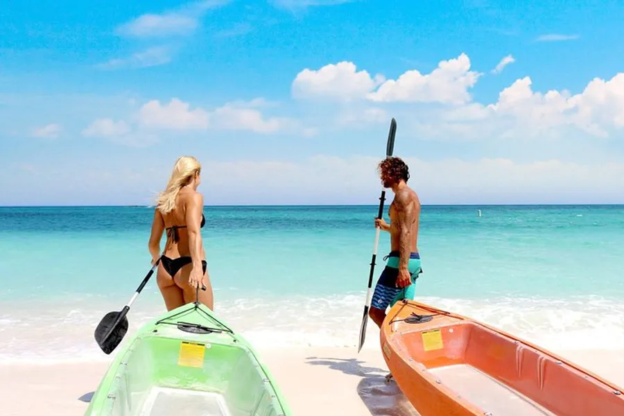 A man and a woman are preparing colorful kayaks on a sunny beach with clear turquoise waters.