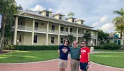 Three individuals are posing for a photo in front of a large two-story house with a green lawn and palm trees under a bright sky.