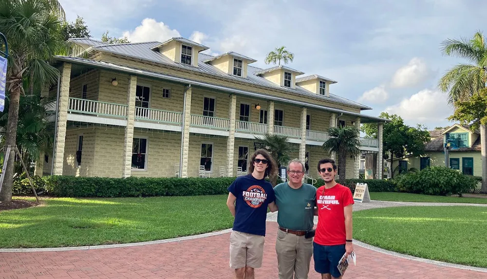 Three individuals are posing for a photo in front of a large two-story house with a green lawn and palm trees under a bright sky