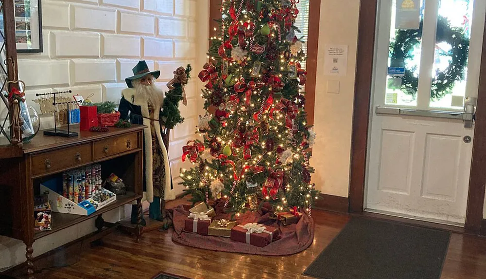 A festively decorated Christmas tree with red accents stands next to a life-size figure dressed as a Christmas character in an indoor setting