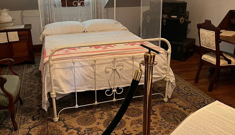 The image shows a vintage-style bedroom with an ornate white metal bed patterned bedding lace-trimmed pillowcases traditional furniture and a protective barrier suggesting this is a display in a museum or historic house