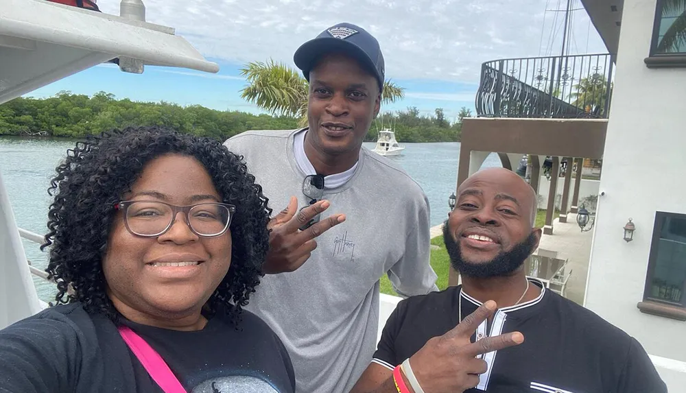 Three people are posing for a selfie with two of them making peace signs in front of a waterfront setting with boats and a building in the background