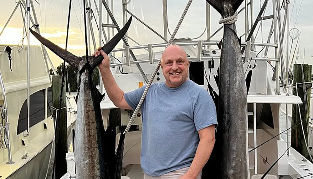 A person is smiling while posing with a large fish possibly a swordfish or marlin hung upside down by its tail at a marina with boats in the background