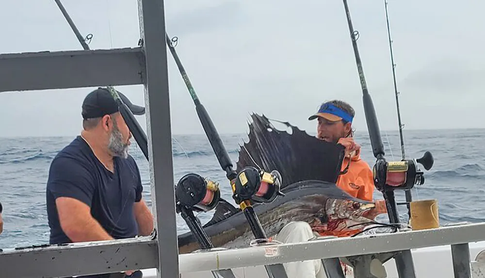 Two people are on a boat with one holding a large recently caught sailfish next to some fishing rods